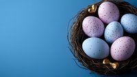 Pastel blue and pink speckled Easter eggs arranged in natural twig nest with golden ribbon accents against vibrant blue background, festive spring holiday concept.