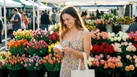 Woman with long brown hair wearing a floral dress shopping at outdoor flower market surrounded by colorful tulips and blooms, holding shopping bag.