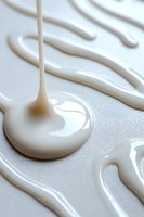 Flowing white liquid drops and streams creating abstract patterns on light surface, macro photography showing viscous texture and glossy finish.