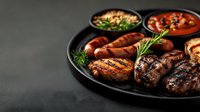 Grilled meat assortment with sausages, pork steaks and chicken served on black plate with fresh herbs and sauces. Dark moody background for menu.
