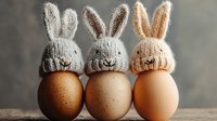 Easter eggs decorated with knitted bunny hats in white, gray and beige colors arranged in row on wooden surface against blurred background. Cozy holiday composition.