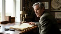 Senior man with gray hair and light complexion, wearing olive green cardigan, seated at wooden desk writing in journal with fountain pen. Warm study interior with candlestick lamp, vintage maps, and natural window light. Suitable for education, literature, memoir, and professional content.