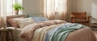 Cozy bedroom with layered bedding in warm earth tones, potted plant on windowsill, natural sunlight streaming through sheer curtains, and leather armchair in corner for relaxation and home decor.