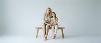Mother and daughter in matching beige outfits sitting together on wooden bench against white studio background for family portrait photography.