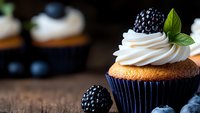 Fresh vanilla cupcake topped with swirled cream frosting, ripe blackberry and mint leaf garnish on rustic wooden background, selective focus macro shot.