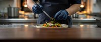 Chef in black uniform and gloves plating gourmet dish with tweezers on dark counter in professional kitchen with warm lighting.