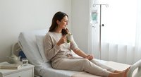 Young woman patient in beige pajamas drinking tea while resting in modern hospital bed with IV drip stand, recovery and healthcare comfort concept.