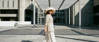 Woman in cream linen suit and straw hat walking through modern urban plaza with geometric architecture and strong shadows.