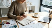 Young woman writing in notebook at modern workspace with laptop and coffee cup for business planning and productivity.