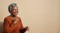 Senior African American woman with gray hair wearing rust-colored ribbed sweater, smiling warmly while gesturing with hand against neutral beige background, expressing joy and confidence.