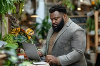 African American businessman working on laptop in modern plant-filled cafe or coworking space with natural lighting and greenery background.