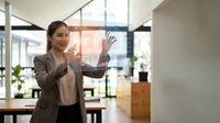 Professional businesswoman presenting data on transparent digital screen in modern office environment with natural lighting and contemporary workspace.