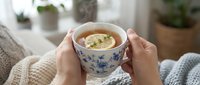 Woman holding decorative mug with hot beverage and lemon slice, wearing cozy sweater indoors near potted plants, perfect for wellness and lifestyle content.