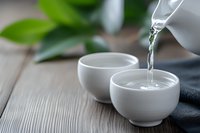 Pouring hot water from white ceramic teapot into small cups on wooden table with green leaves in background for tea ceremony and relaxation moments.