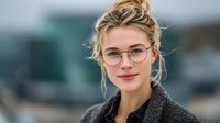 Young blonde Caucasian businesswoman with glasses and messy bun hairstyle smiling confidently in modern office environment for corporate presentations.