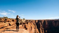 Hiker with backpack standing on rocky cliff edge overlooking vast canyon landscape under clear blue sky, adventure travel and outdoor exploration concept.