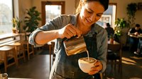 Smiling female barista in apron pouring steamed milk into a latte cup at a modern coffee shop, creating latte art with natural light.