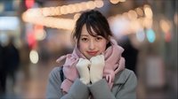 Young Asian woman in pink hoodie and gray jacket smiling warmly at camera with hands near face on blurred city street background with bokeh lights.