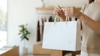 Woman in beige linen shirt holding blank white shopping bag with handles in modern home interior with plant and clothing rack.