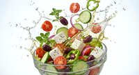 Fresh Greek salad ingredients splashing in glass bowl with tomatoes, cucumber, olives, feta cheese and herbs on white background.