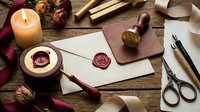 Vintage wax seal stamp set with envelope, candle, dried roses, scissors, and ribbon on rustic wooden table for elegant correspondence.