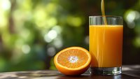 Fresh orange juice being poured into glass with orange half on wooden table against blurred green nature background for healthy breakfast concept.