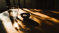 Glass of water with dramatic sunlight and shadows on wooden table, small dark bowl with salt creates minimalist composition in moody lighting atmosphere.