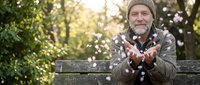 Senior Caucasian man with beard sitting on park bench blowing flower petals from hands during golden hour sunset creating magical moment in nature.