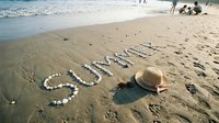 Word summer written with seashells on sandy beach, straw hat and sunglasses nearby, ocean waves in background, sunny vacation mood.