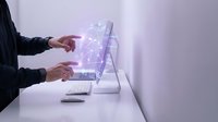 Businesswoman in dark blazer interacting with holographic digital interface on desktop monitor, displaying glowing purple data visualization and network connections for technology and innovation concepts.