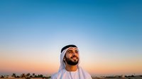 Middle Eastern man wearing traditional white headwear and thobe against desert sunset sky with palm trees. Suitable for cultural representation, travel, lifestyle, and business content.