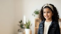 Young girl with curly dark hair and medium skin tone wearing denim vest over striped shirt, smiling at camera in bright indoor space with potted plants and natural light.