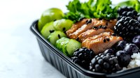 Fresh salmon poke bowl with avocado, blackberries, rice and sesame seeds on white marble background, healthy lunch meal prep in black container.