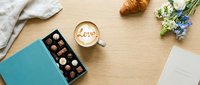 Flat lay of cappuccino with love latte art, open chocolate box, croissant, and fresh flowers on wooden table for romantic morning concept.