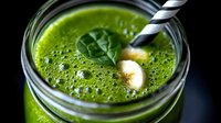 Fresh green smoothie with spinach leaves and banana in glass jar with striped straw, healthy breakfast drink closeup on dark background.