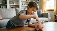 Young boy in striped shirt putting coins into pink piggy bank at home, learning about saving money and financial literacy for children.