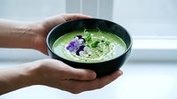 Hands holding black bowl with creamy green soup garnished with purple flowers and microgreens, ready for serving or food photography.