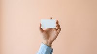 Close-up of a hand holding a blank white card against a neutral beige background, suitable for business, marketing, and promotional designs.