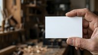 Male hand holding blank white business card in carpentry workshop with tools and wood shavings in background. Suitable for branding, business identity, and craft trade marketing.