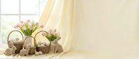 Easter bunny rabbits with wicker baskets and spring tulip flowers on wooden table by sunny window with yellow curtains for holiday decoration.