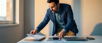 Professional businessman in navy suit reviewing documents at modern office desk during golden hour lighting for corporate presentations and business strategy meetings.