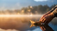 Hand placing paper boat on calm water surface during golden hour sunset with warm bokeh lights reflecting on lake creating peaceful moment.