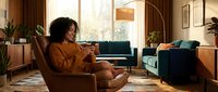 Young woman with curly dark hair, wearing a mustard-colored sweater, relaxing in a mid-century modern living room with vintage furniture and warm lighting.