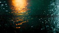 Dramatic sunset reflection on wet asphalt surface with golden bokeh lights and water droplets creating moody atmospheric urban scene in dark teal tones.