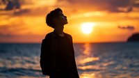 Young Asian man silhouette against dramatic orange sunset sky over ocean, looking up with hopeful expression. Peaceful serene moment captures aspirational mood.