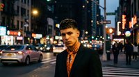 Young Hispanic man in black suit standing on busy city street at night with glowing lights and traffic creating urban atmosphere for lifestyle content.