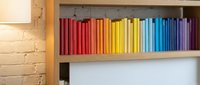 Colorful rainbow books arranged on wooden shelf creating vibrant spectrum from red to purple against white brick wall for home library organization.