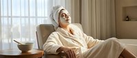 Young woman in white bathrobe and towel relaxing in spa chair with face mask, enjoying wellness treatment in luxury hotel room with natural lighting.