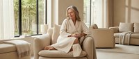 Mature woman with blonde hair, Caucasian ethnicity, wearing cream cardigan, relaxing in modern living room with neutral tones, natural light from windows, contemporary furniture and decor for lifestyle and wellness content.