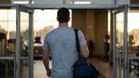 Young man with backpack walking through glass doors entering modern building lobby during golden hour sunset lighting for education and lifestyle concepts.